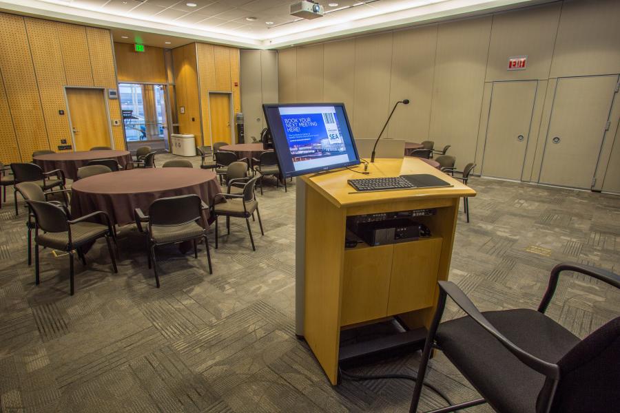 View from the podium located inside the London Conference room featuring 6 banquet round tables with brown table clothes and chairs.